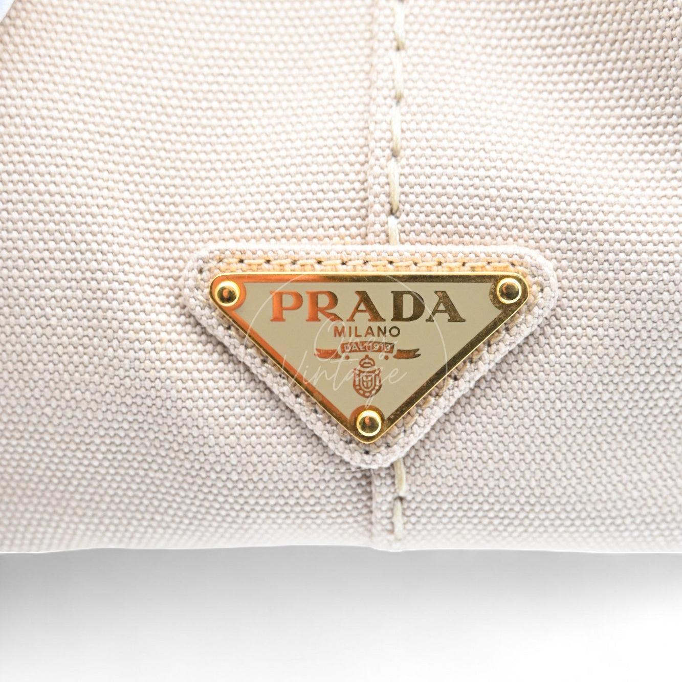 Close-up of Prada gold-tone metal triangle logo badge on beige canvas bag